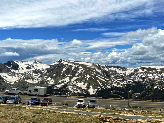 Summer views in the Rocky Mountains. Image shows view from near Alpine Visitor Center off Trail Ridge Road. Many cars and vans off a high mountain road with snow covered mountains in the background.