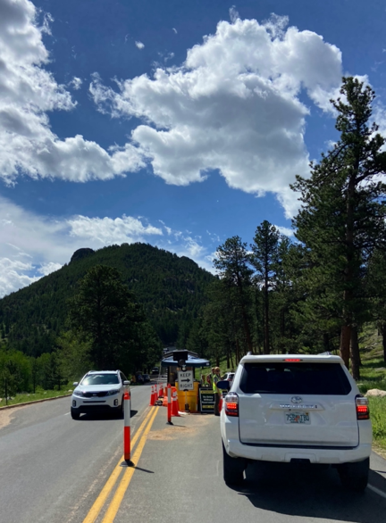 Busy entrance at Bear Lake Corridor during a summer trip at Rocky Mountain national park. Image shows a line of cars entering a hilly green valley.