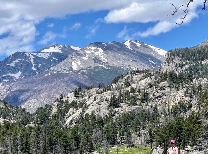 image shows a woman in a pink hat, pink shirt with pink hair standing on a boulder with the rockies in the background.