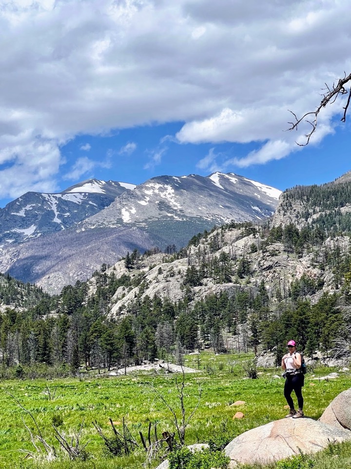 image shows a woman in a pink hat, pink shirt with pink hair standing on a boulder with the rockies in the background.