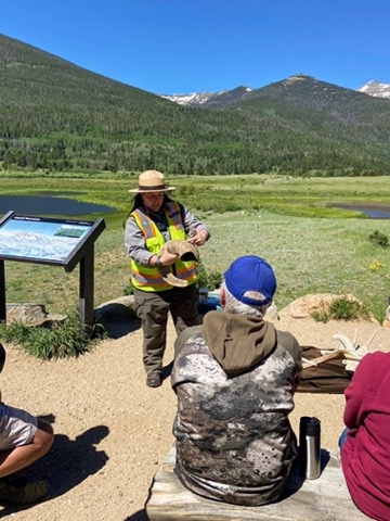image shows a National Park ranger holding a bighorn sheep horn at Rocky Mountain National Park, while giving a lecture on bighorn sheep. Behind is a lake and mountain.