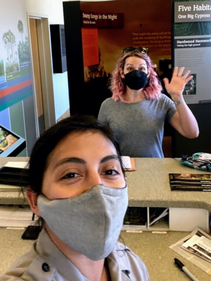 image shows a brown haired woman woman in a face mask taking a selfie with a woman with pink hair at a ranger station
