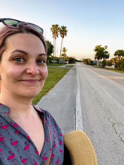 image shows a woman with pink hair and a flamingo shirt taking a selfie in front of a very empty road in Everglades City