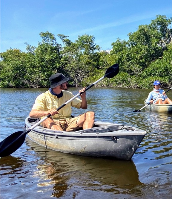 image shows a man with a black bucket hat and yellow shirt in a gray canoe in water with mangrove trees in Fakahatchee Strand