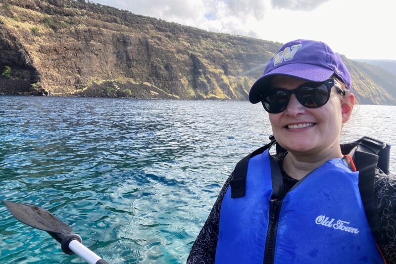 image shows a woman kayaking in a purple hat and blue life jacket in blue water