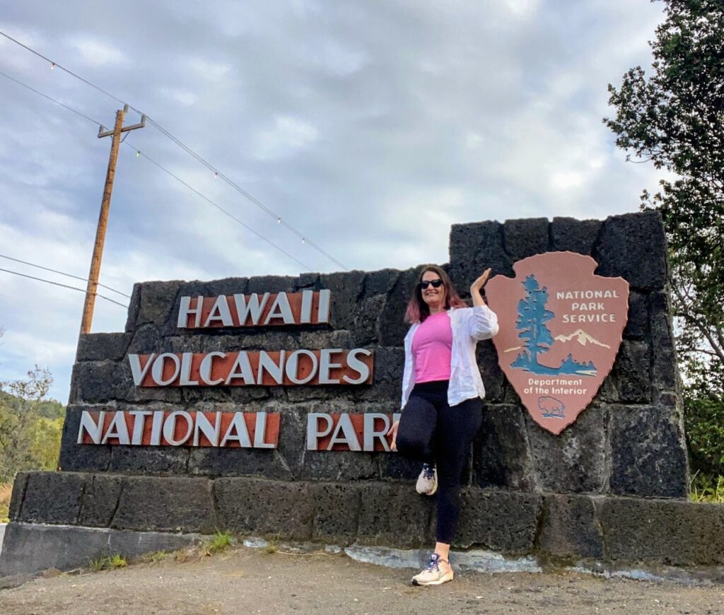 image shows a woman in a pink shirt in front of a sign for Hawaii Volcanoes National Park