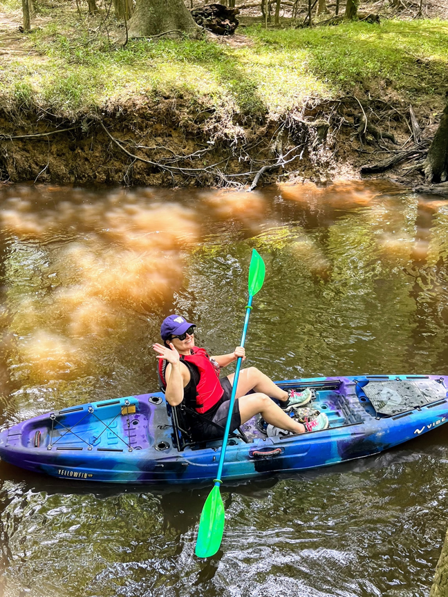 woman kayaking in Cedar Creek in a blue kayak with red vest