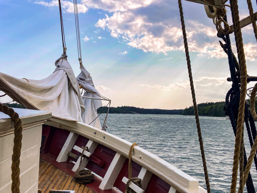 image shows the bow of a wooden sailboat at sunrise, with rays coming through the clouds