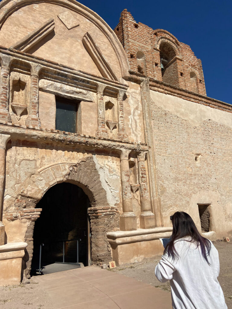 image shows a large Spanish mission at Tumacacori with a woman in a white sweater in front
