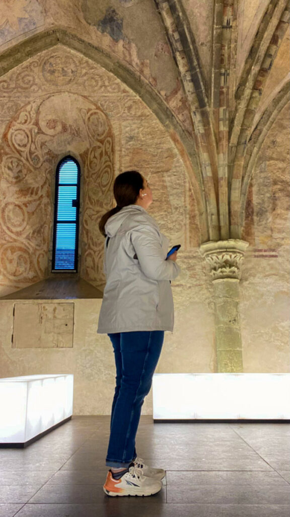 image shows a woman in a white jacket and jeans listening to an audio guide while at chateau de chillon, in a chapel.