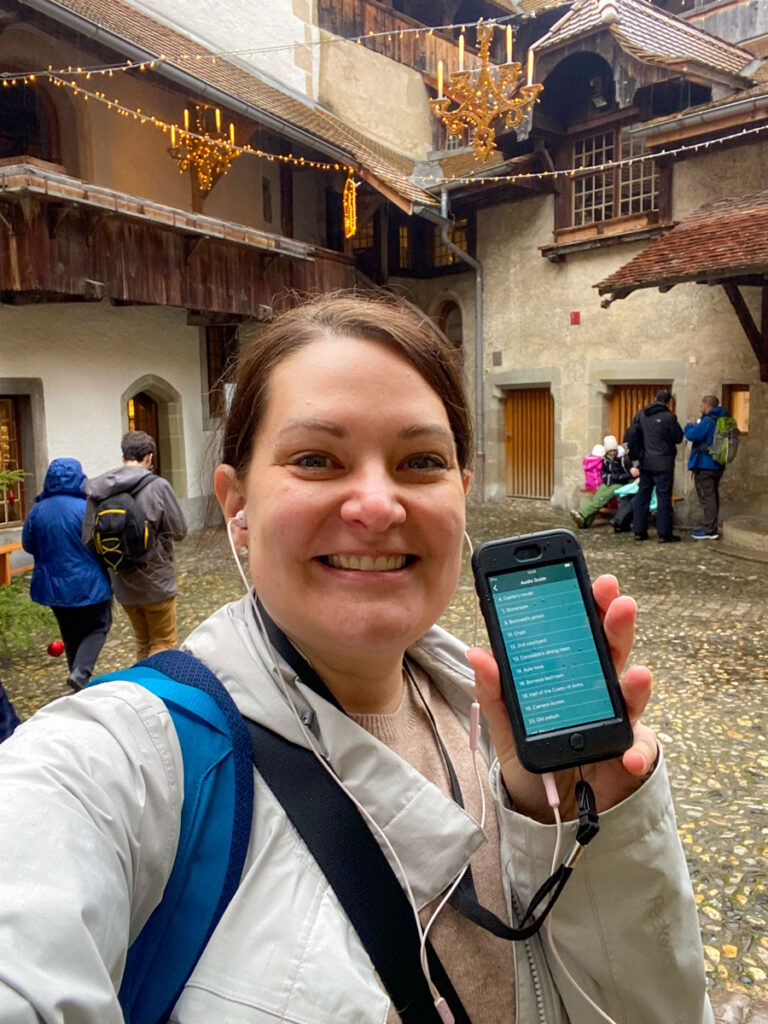 a woman smiles with pride as she holds up an old iphone with an audio guide onit. she is standing in the middle of a chateau keep in the rain on a wintery day in montreux