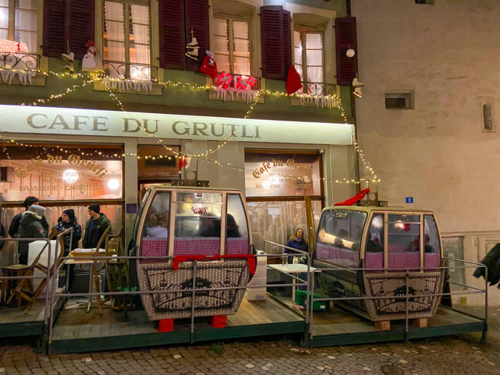 two ski gondola cars in front of a restaurant decked for christmas. people are dining inside the small cramped ski cars in lausanne