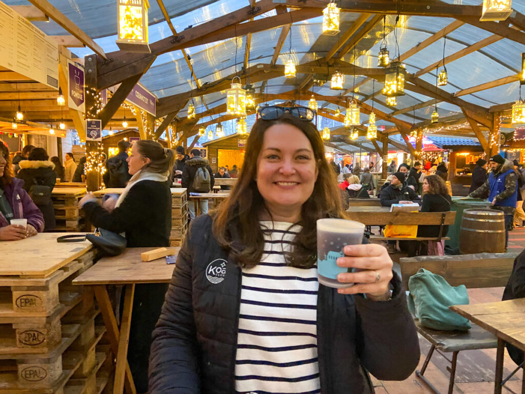 image shows a woman in a black coat with a striped shirt holding a glass of mulled wine, in a hall at a christmas market in Lausanne in winter