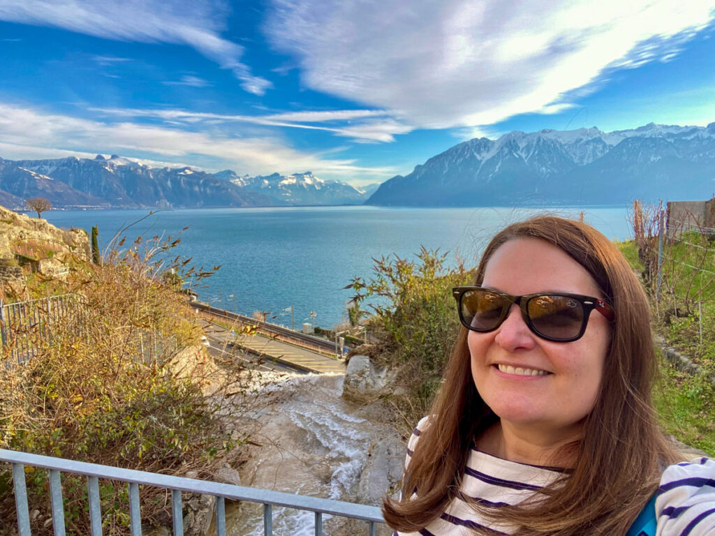 a woman with brown hair and sunglasses in a striped shirt poses for a selfie in front of lake geneva. she is not wearing a coat even on this winter visit because it is sunny