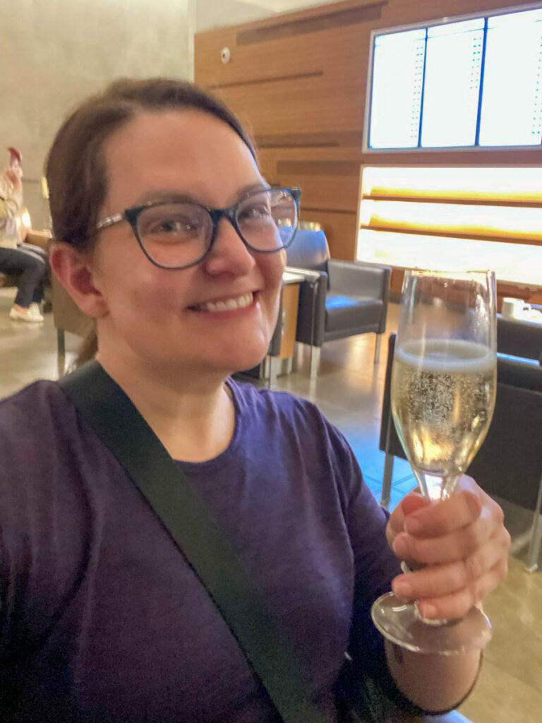 a slightly blurry image shows a woman in a purple shirt holding a glass of champagne in an airline lounge