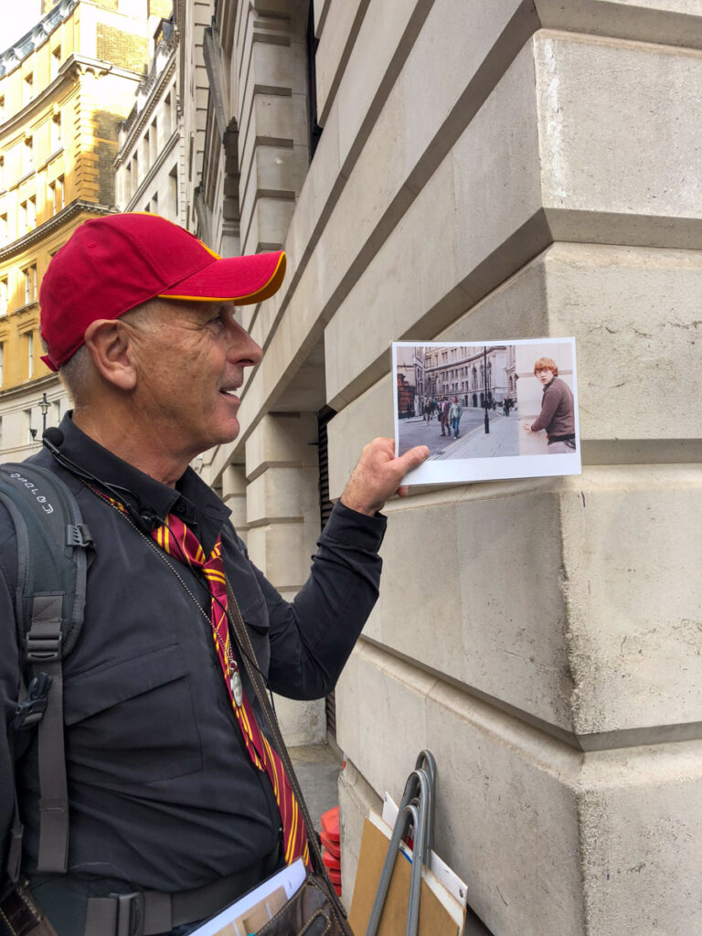 image shows a British man in a red hat holding up a still from a Harry Potter movie where it took place.