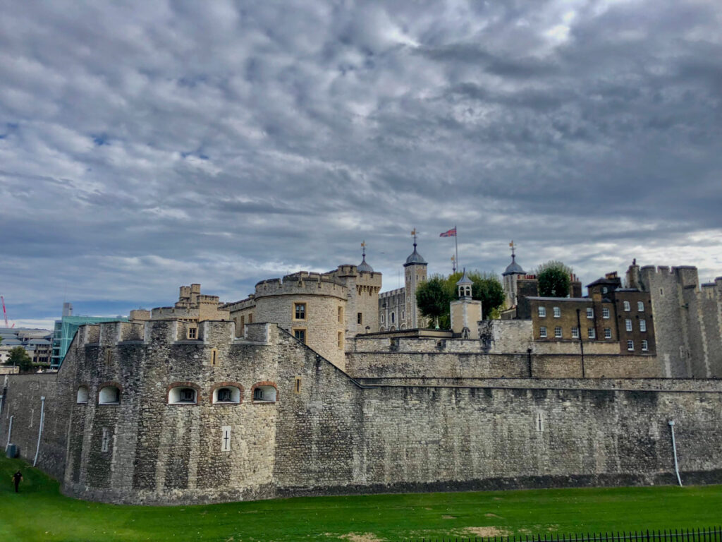 image shows the Tower of London gray stone outer wall surrounded by the green of a moat and a gray sky.