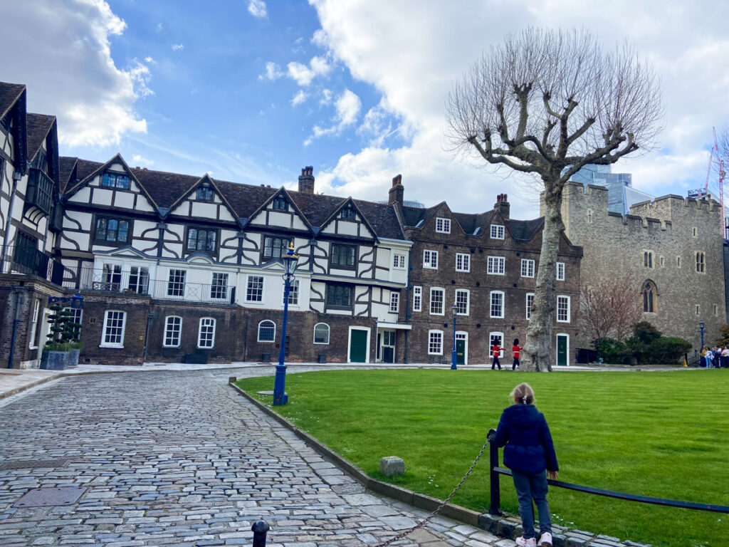 image shows the tudor homes of the Tower of London surrounding a green courtyard. a small girl looks on at soldiers in the distance