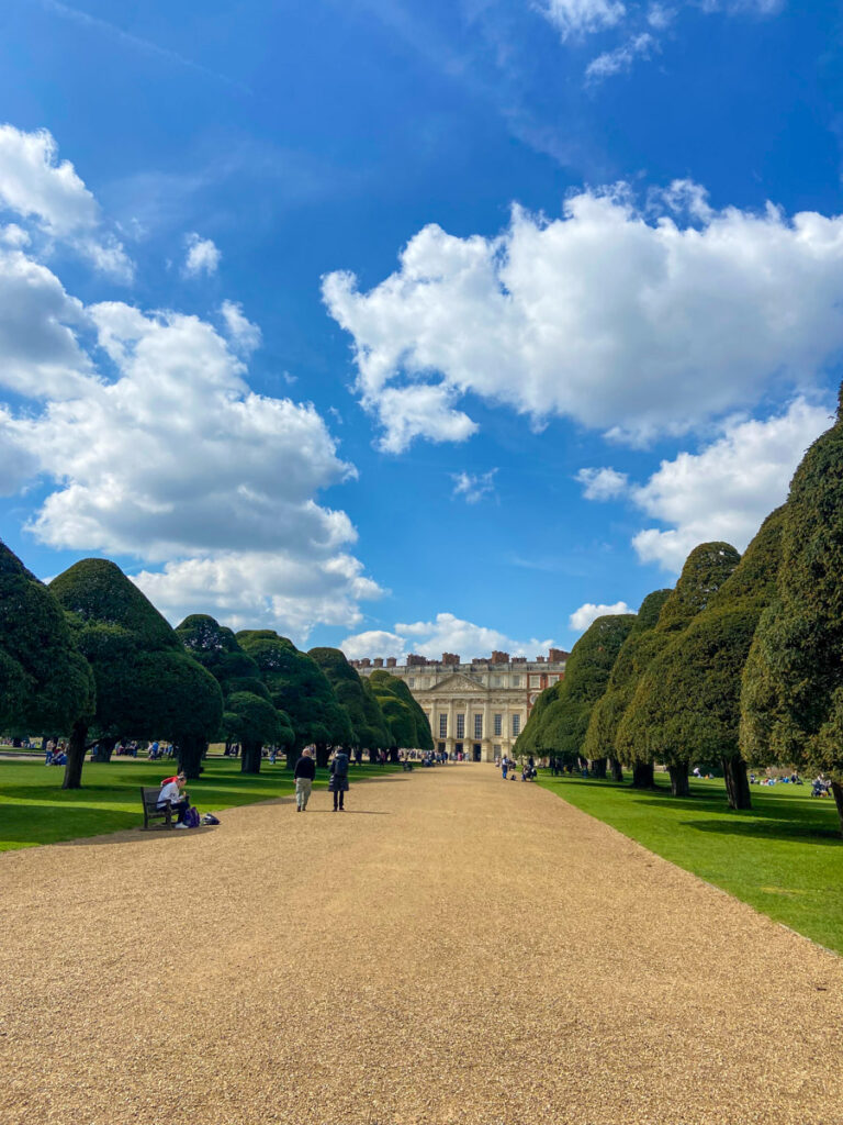 image shows a wide gravel path, with perfectly trimmed yew trees on either side, leading to a palace at the end