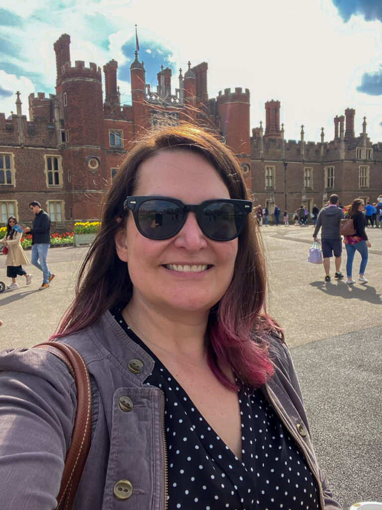 image shows a woman in a black top with a purple jacket and sunglasses on in front of the red brick of Hampton Court Palace in the UK