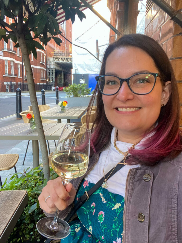 a woman with brown and pink hair holding a glass of white wine smiling at a bistro in london