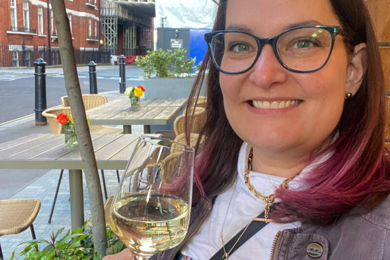 a woman with brown and pink hair holding a glass of white wine smiling at a bistro in london