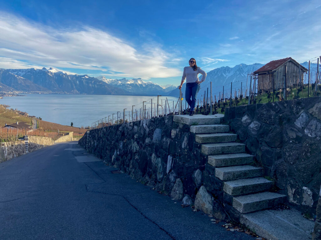 a woman in sunglasses and s triped shirt poses for a photo at the top of small concrete steps on one of many hiking trails in lavaux winery region in the winter