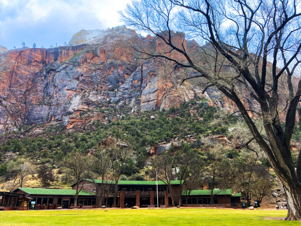 the Zion Lodge is a rustic wooden and stone structure with a green roof. a large green lawn sits out front. Behind it is the imposing rock of the Zion canyon.