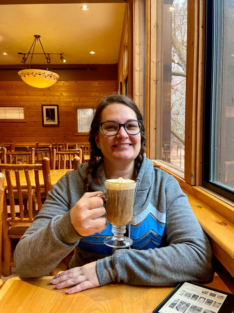 woman with brown hair in braids and a gray sweatshirt sits in a rustic lodge drinking a boozy coffee drink
