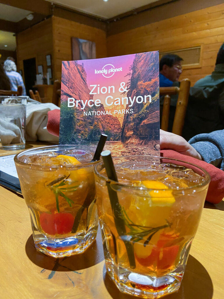 a Lonely Plane book on Zion National Park stands propped up in front of two alcoholic drinks in rocks glasses, with an orange, cherry and rosemary garnish. The setting is a rustic lodge.