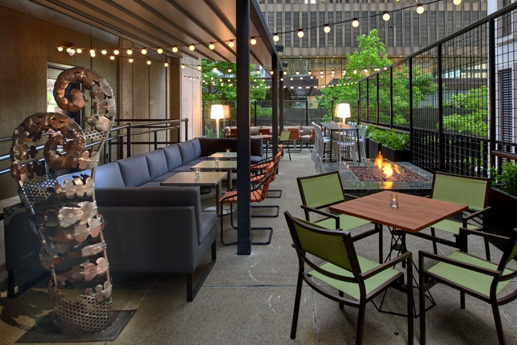 an outdoor hotel patio with a long blue banquette sofa with small tables and chairs, plus a small patio table and four green chairs in the foreground plus a firepit