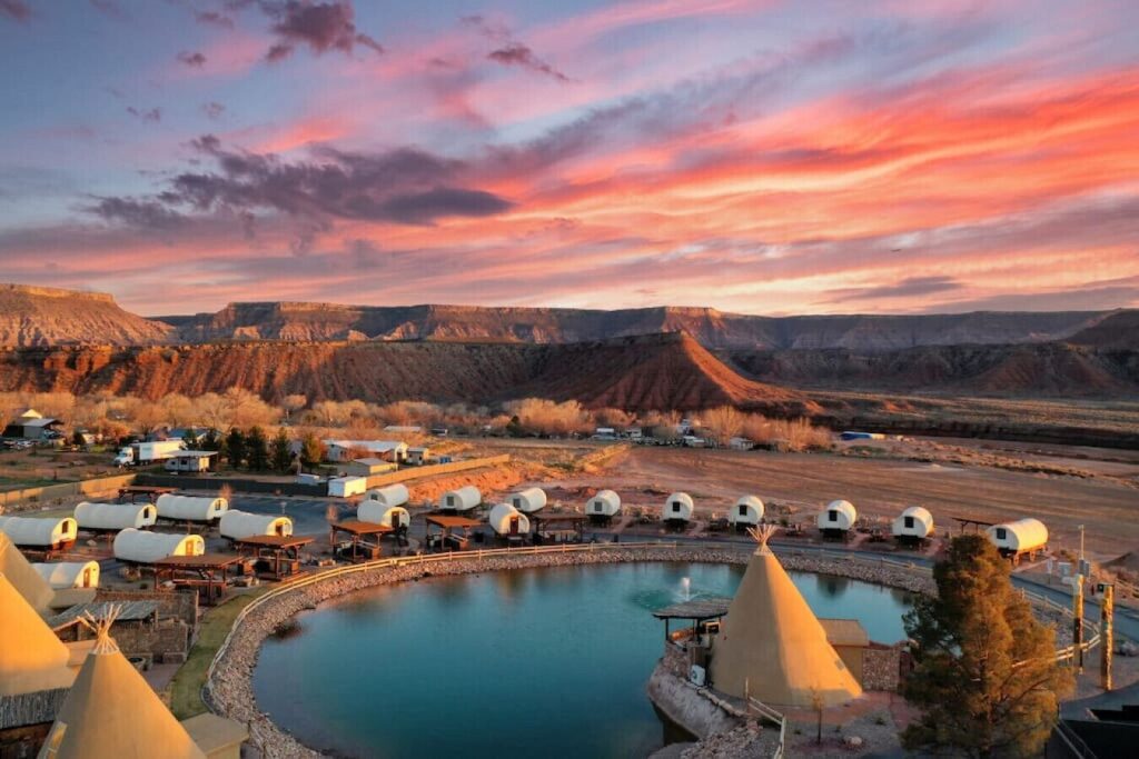 image shows several old covered wagons that are now hotel rooms around a lake with canyons in the background.