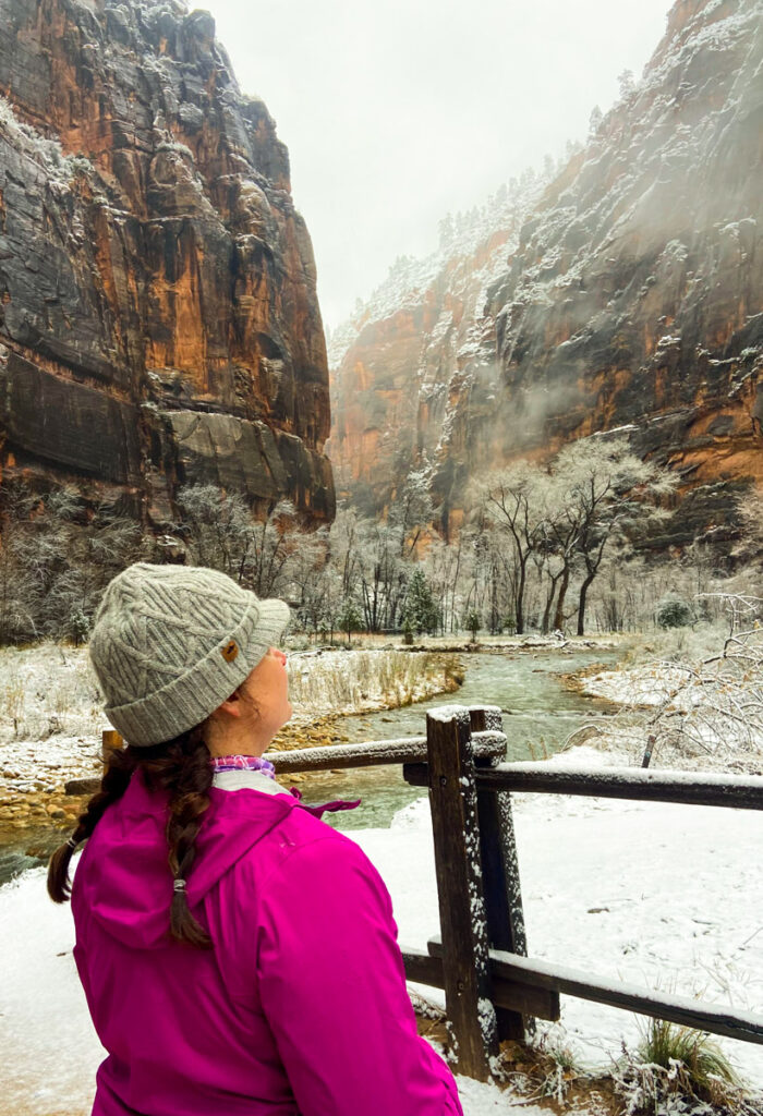 A woman in a bright pink rain jacket, gray winter hat and braids looks out over the Virgin River and the snow covered banks in the deep canyon