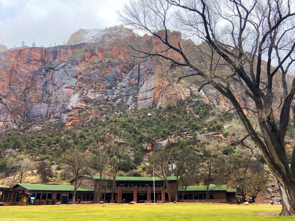 the Zion Lodge is a rustic wooden and stone structure with a green roof. a large green lawn sits out front. Behind it is the imposing rock of the Zion canyon.