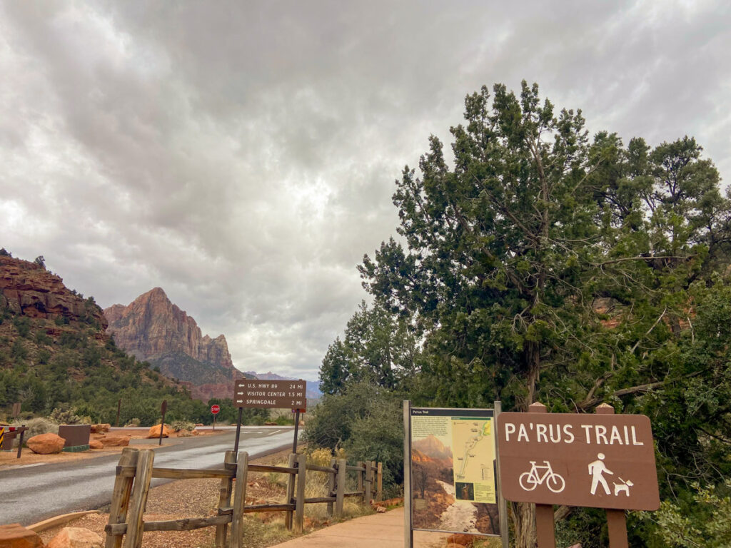 In the foreground a wooden sign for Pa'rus trail in Zion National Park shows it is bike and dog friendly. Behind in the background is the Virgin River and the red rocks the park is known for.